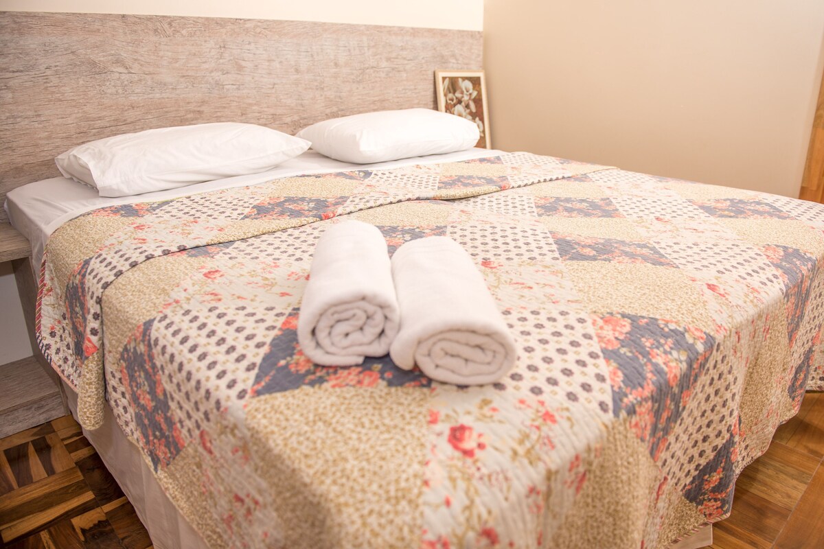 A neatly made bed with a floral-patterned bedspread, two rolled towels, and white pillows.