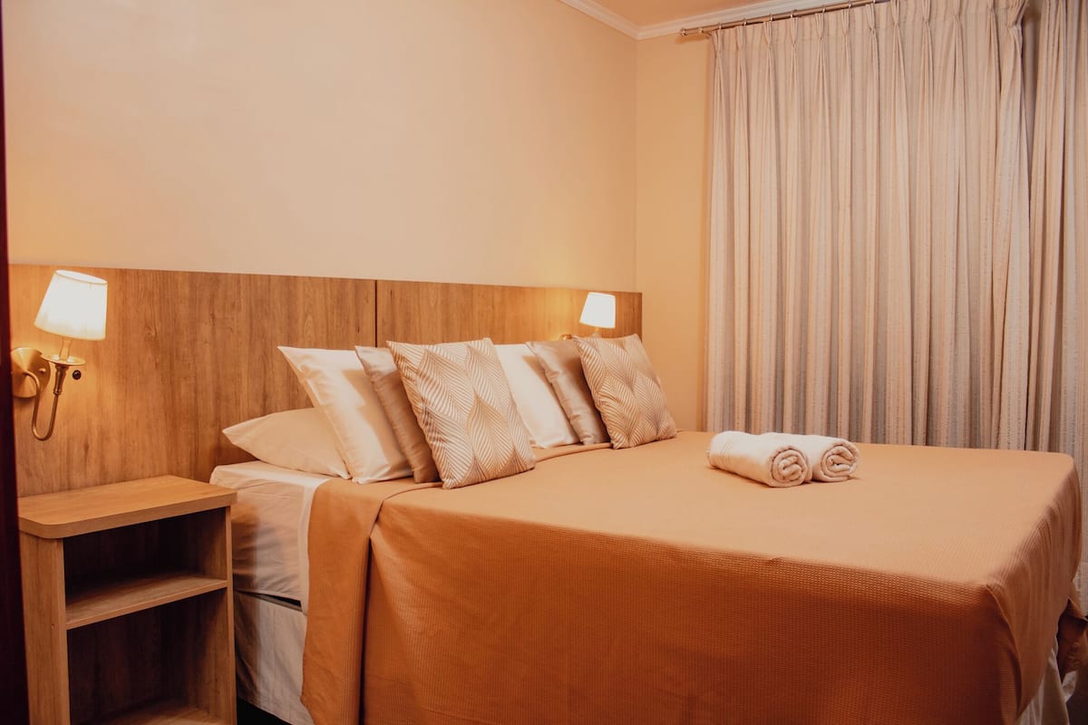 A hotel room with a wooden headboard, a bed with beige bedding, two bedside lamps, and a curtain on the window.