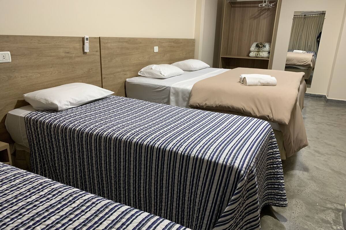 A hotel room with three beds, each with a different patterned bedspread. There is a wooden headboard, a wardrobe, and a mirror on the wall.