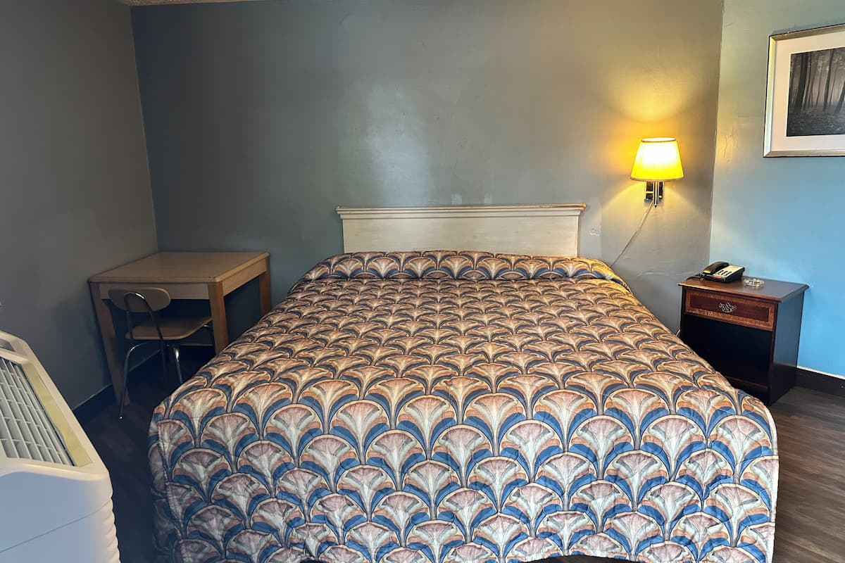 A hotel room with a bed, a desk, a chair, a nightstand, and a lamp.