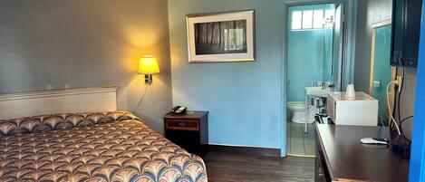 Habitación de hotel con cama, lámpara de noche, mesita de noche, un cuadro, baño con lavamanos e inodoro, y cómoda.