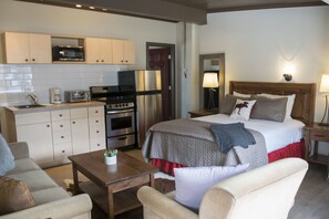 A hotel room with a kitchenette, a bed with multiple pillows, a bedside table with a plant, and a nightstand with a lamp.