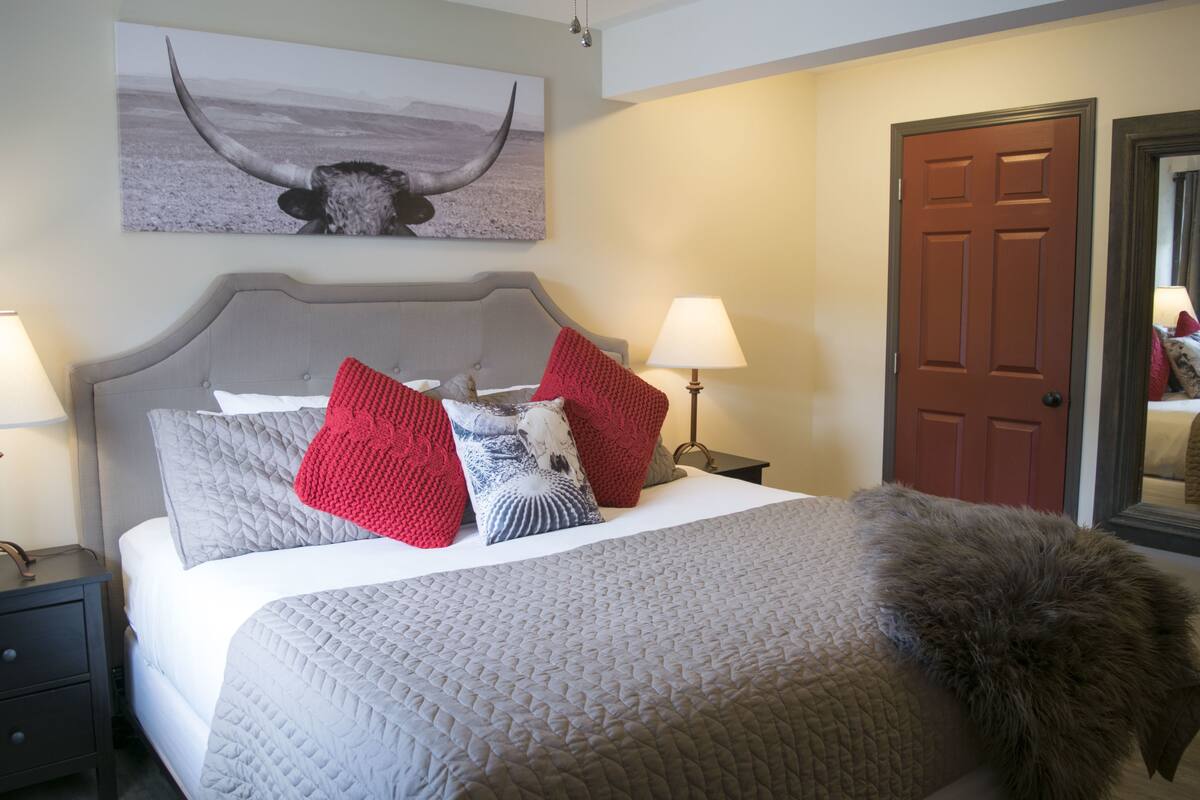 A neatly made bed with a grey quilt, red and white pillows, and a fluffy brown throw.