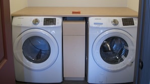 A laundry room with two white Samsung washing machines and a wooden shelf above them. - Yamnuska Suites (Dead Man's Flats)