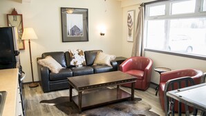 A living room with a leather sofa, a red armchair, a wooden coffee table, and a large window with a view of a car outside. - Yamnuska Suites (Dead Man's Flats)