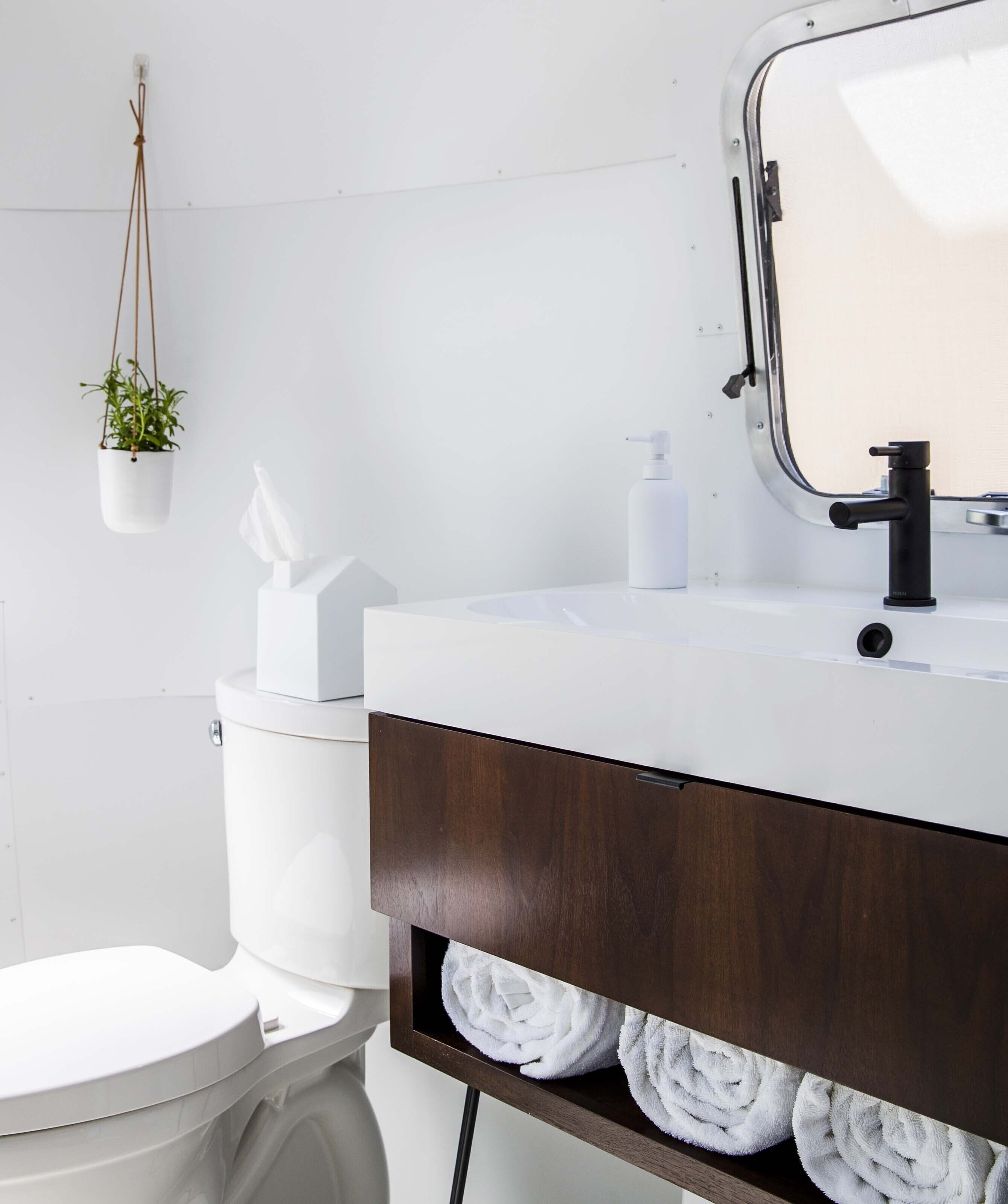 Une salle de bain moderne avec des toilettes blanches, un lavabo blanc doté d’un robinet noir, une plante suspendue et un porte-serviettes avec des serviettes blanches.
