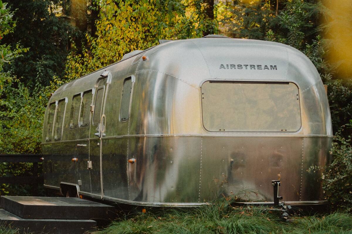 A silver AIRSTREAM trailer with a covered rear section, positioned in a lush garden with a wooden bench and steps leading up to it.