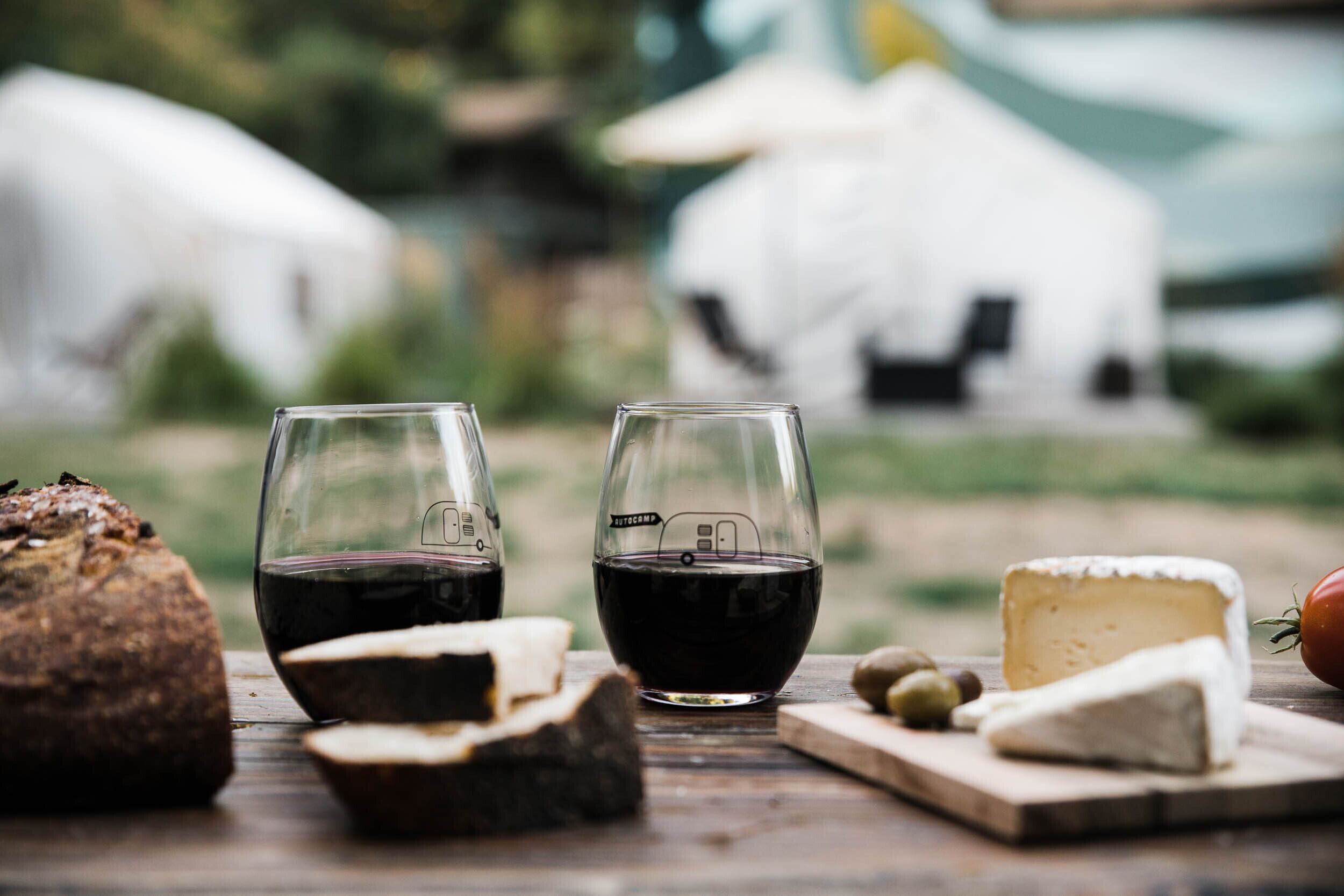 A rustic wooden table with a loaf of bread, two glasses of red wine, a wedge of cheese, and olives.