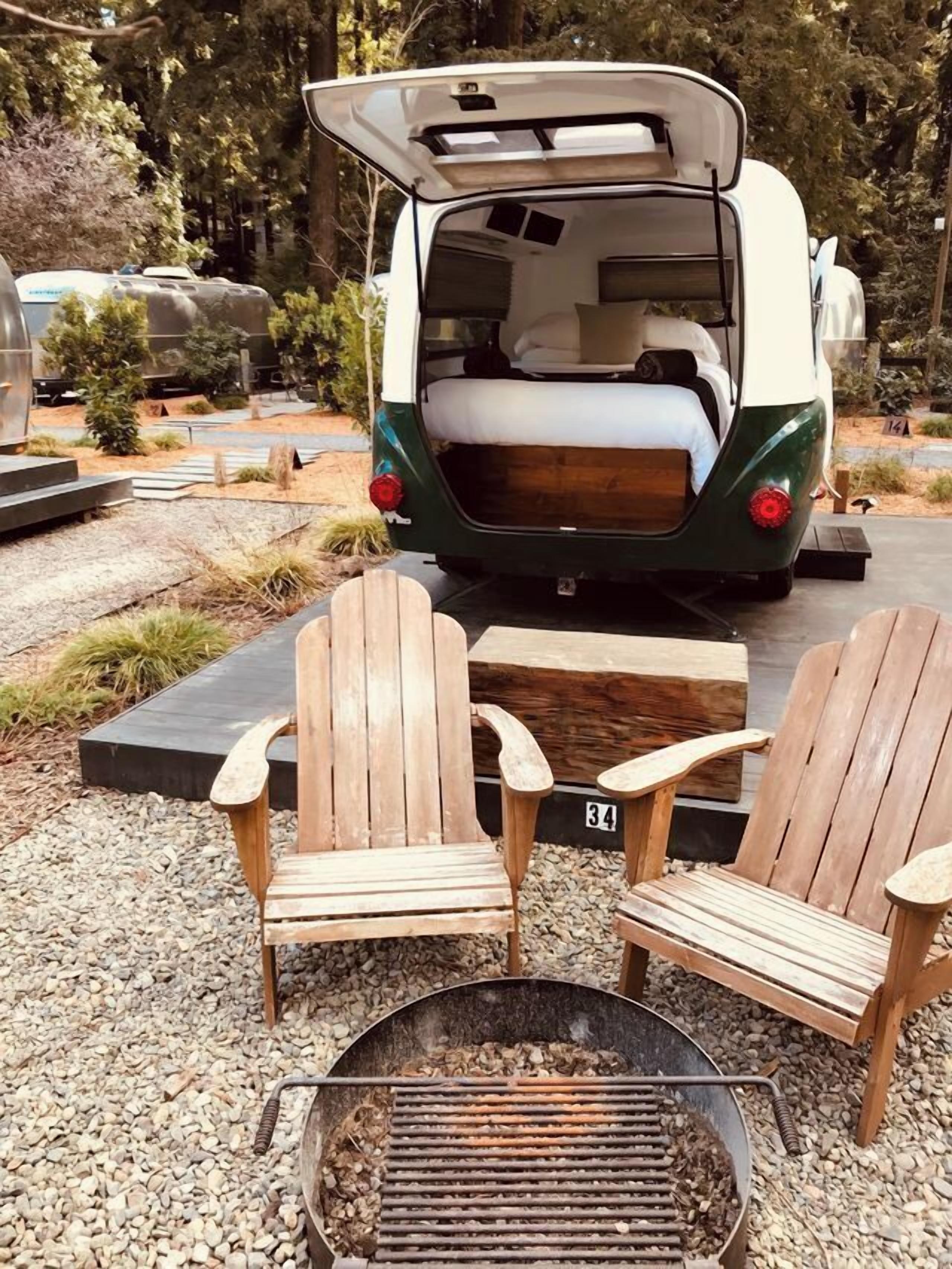 Uma van camper com a parte traseira aberta, duas cadeiras Adirondack de madeira e um braseiro em uma área externa ajardinada.