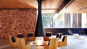 A modern lounge area with a central fire feature, wooden seating, and a backdrop of stacked logs. - AutoCamp Sonoma (Guerneville)