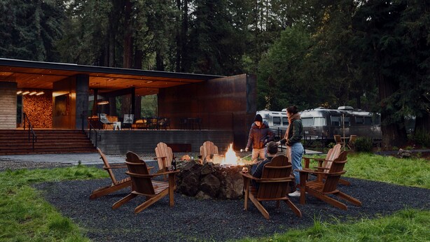 A modern outdoor seating area with Adirondack chairs around a fire pit, a contemporary building with large windows, and a forested backdrop. - AutoCamp Sonoma (Guerneville)