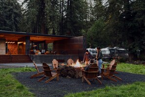 Un espacio al aire libre moderno con sillas Adirondack alrededor de una fogata, un edificio contemporáneo con ventanales amplios y un entorno boscoso.