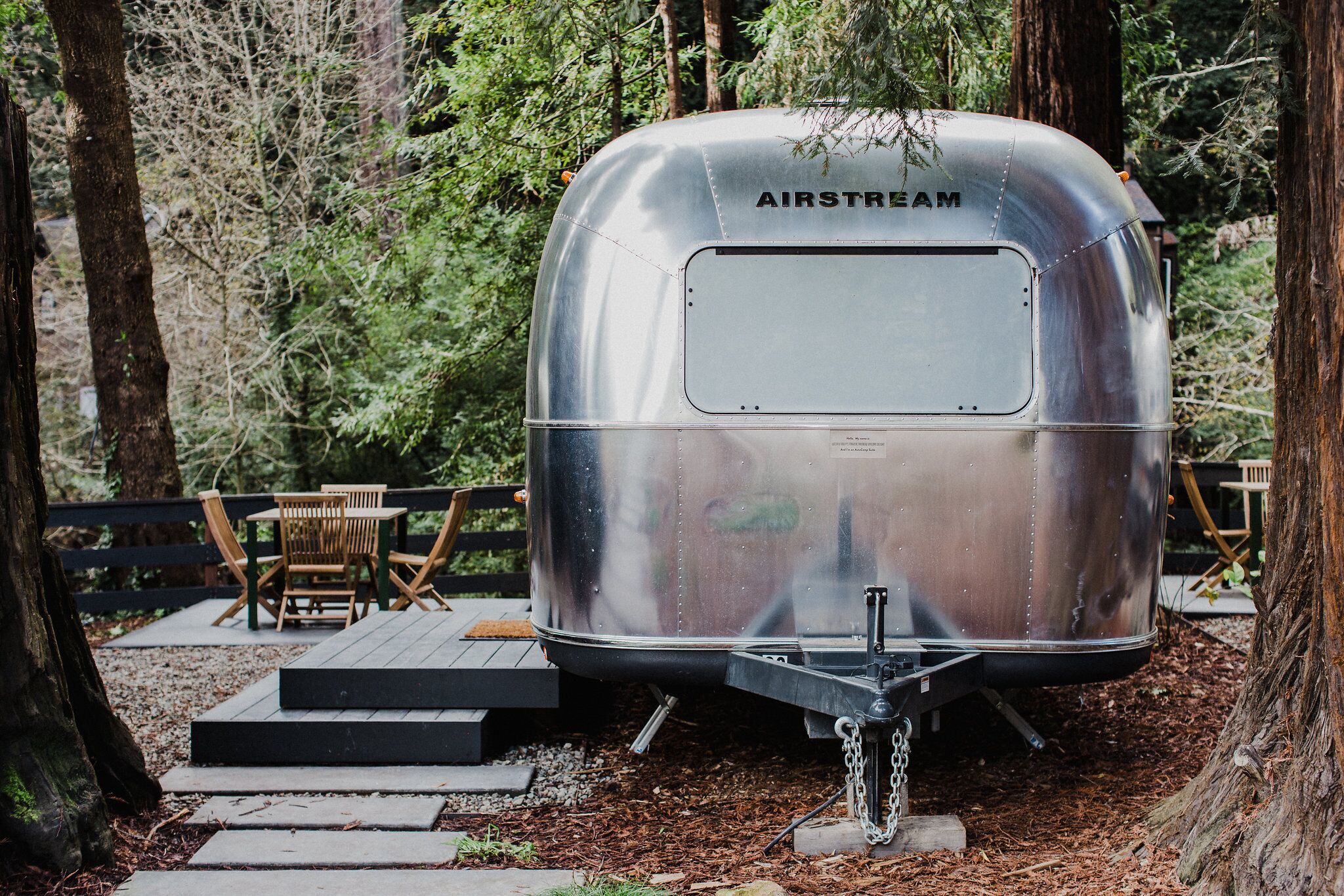 A silver AIRSTREAM trailer with a flatbed, positioned on a wooden deck surrounded by trees.