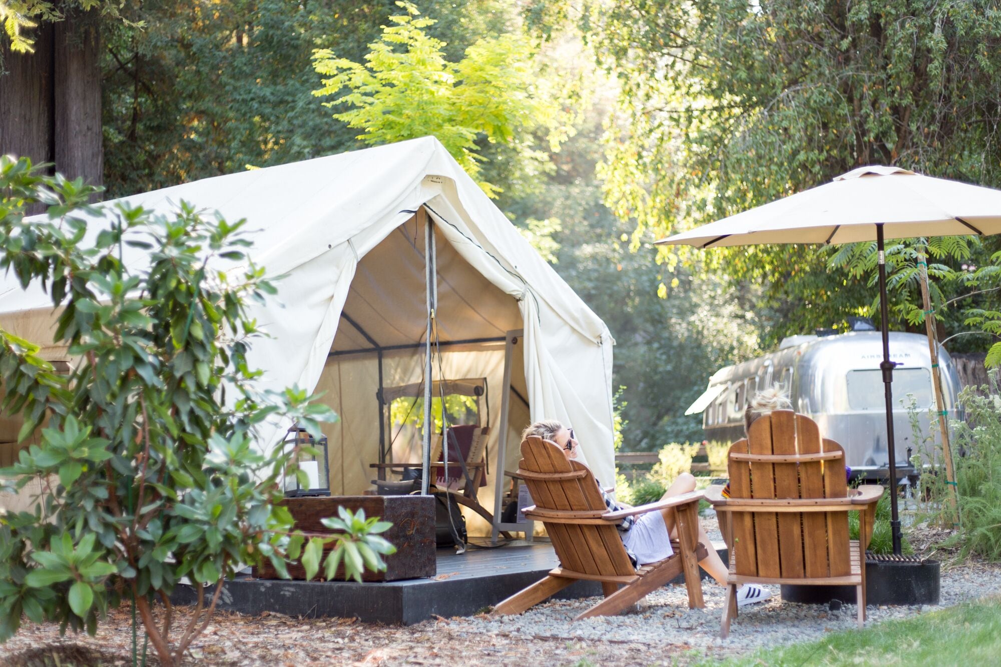 A tented outdoor area with two wooden chairs and a table, surrounded by trees and a camper.