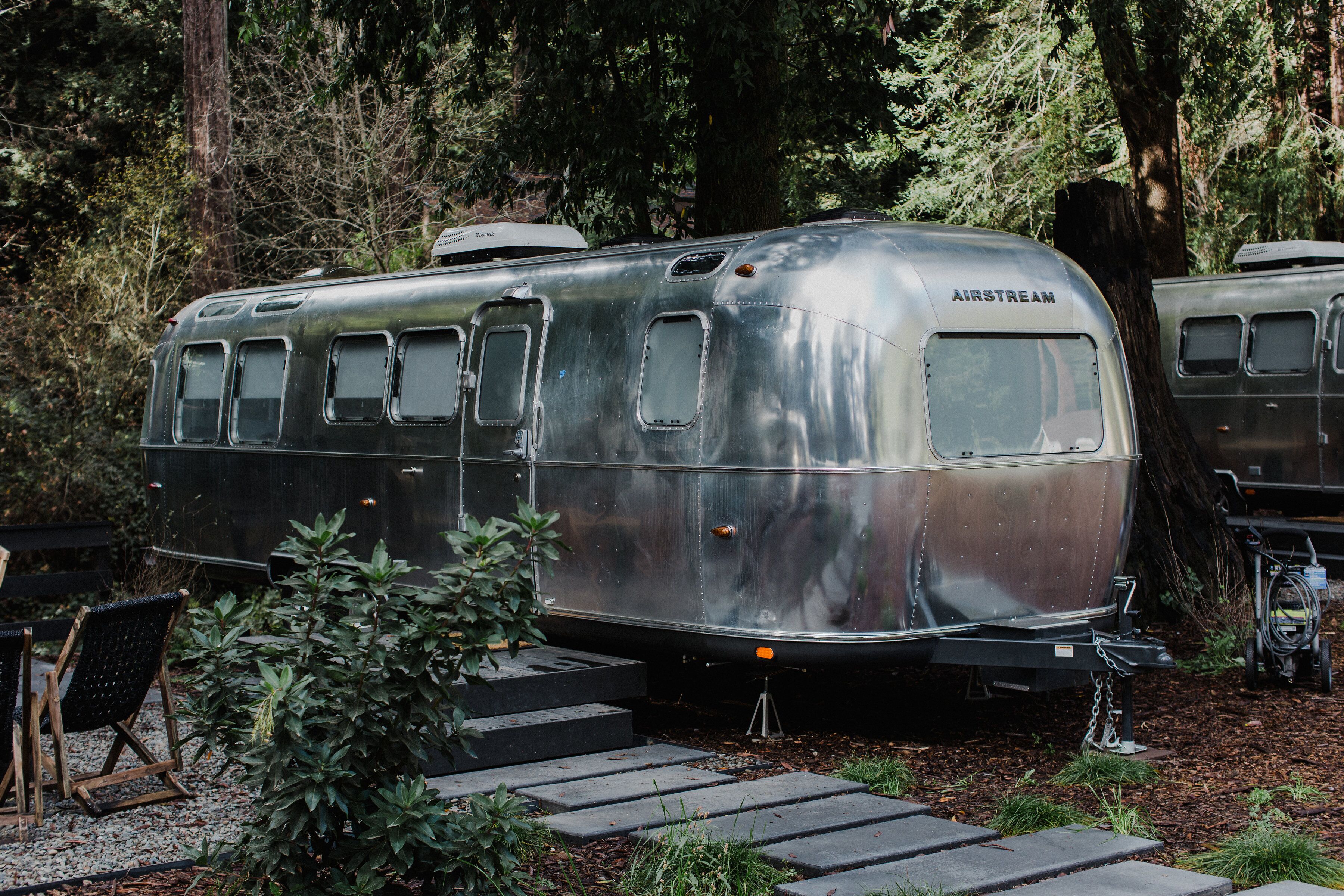 Uma caravana Airstream antiga estacionada em uma área arborizada, com degraus que levam até ela.