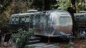 Uma caravana Airstream antiga estacionada em uma área arborizada, com degraus que levam até ela.