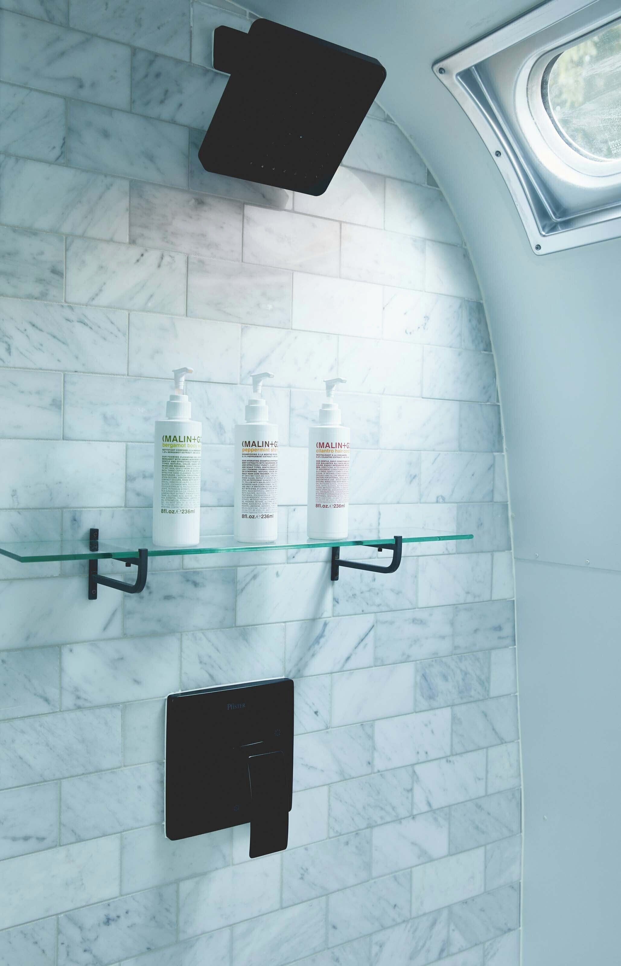 A bathroom with a marble wall, three MALIN+H bottles on a glass shelf, a black towel dispenser, and a window above the shelf.