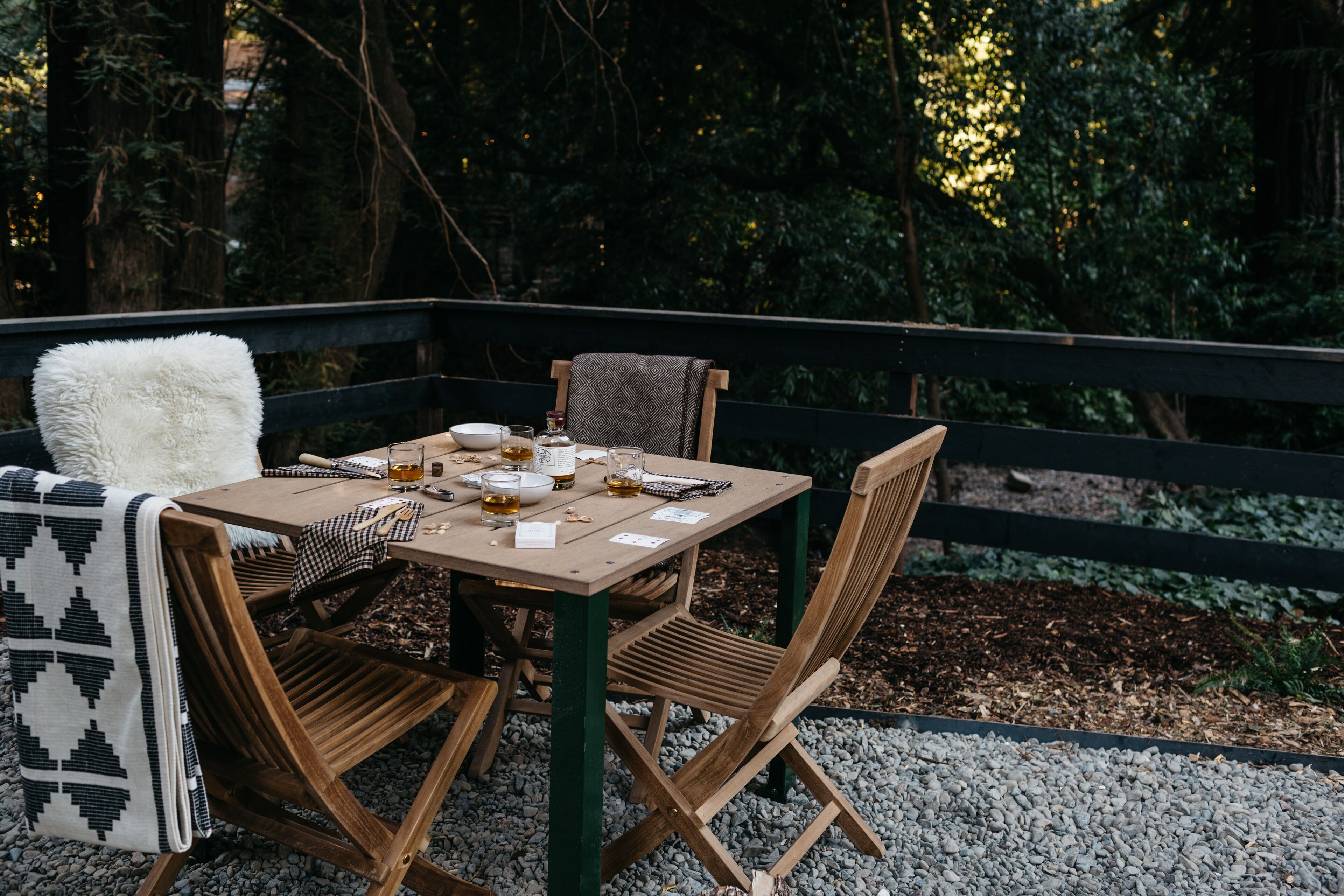 An outdoor dining area with a wooden table and chairs, surrounded by a black railing and a forest backdrop.