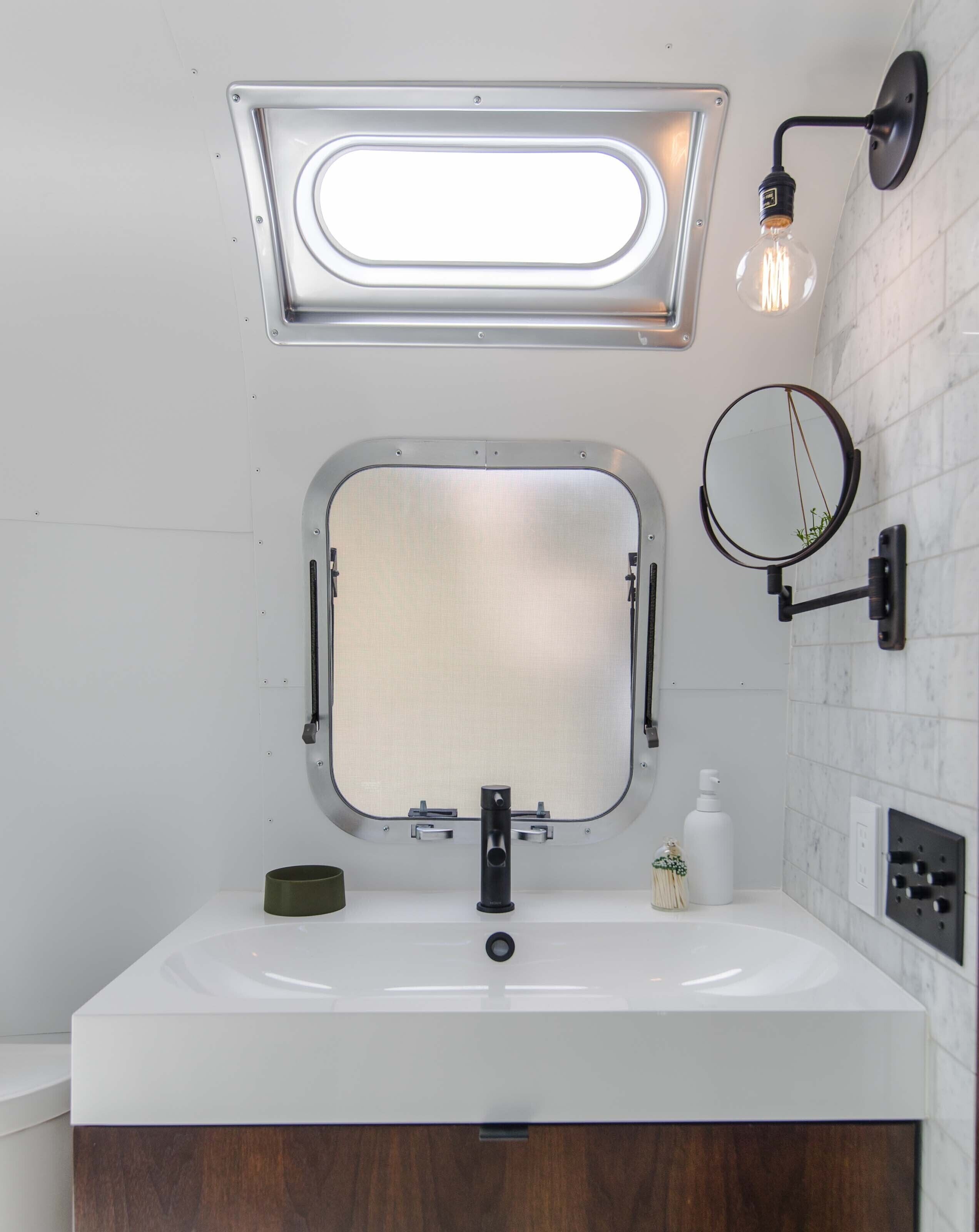A bathroom with a white sink, a mirror, a light fixture, and a small window.