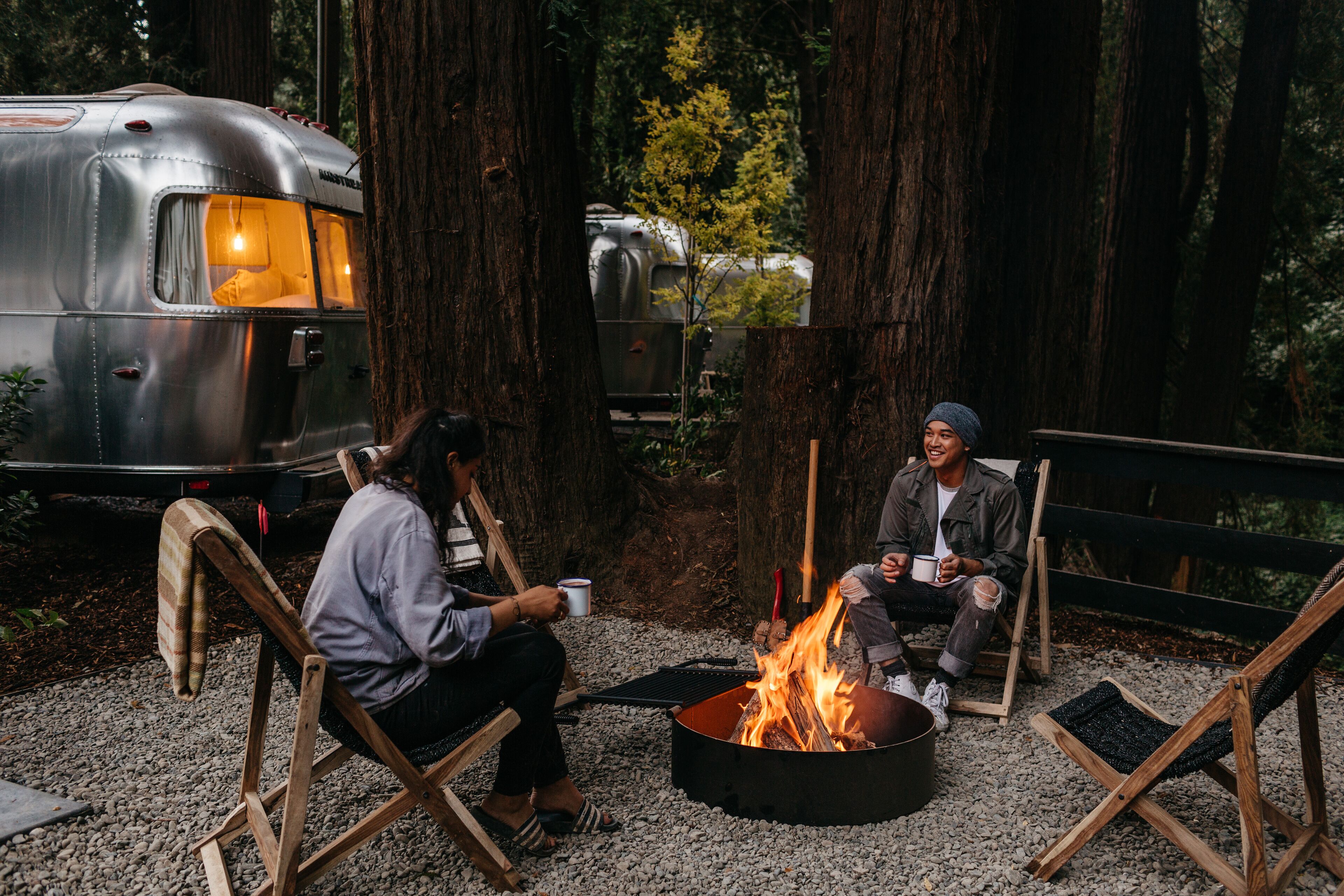 Two people sitting on wooden chairs around a campfire in a forested area with a vintage trailer in the background.