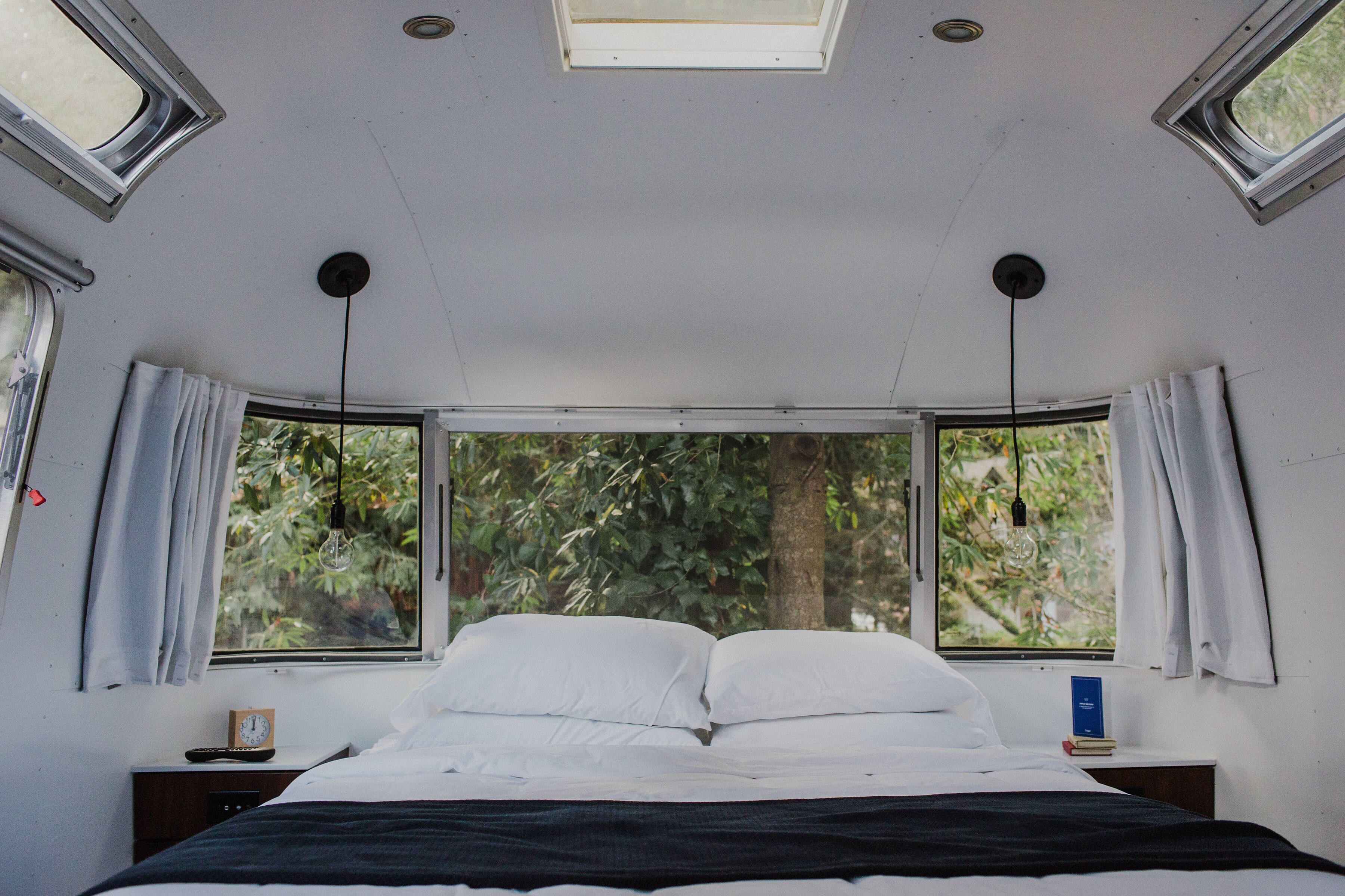 A bedroom with a large bed, two windows, a bedside table with a book, and a clock.