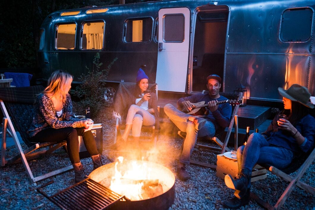 A group of people sitting around a campfire in a trailer with a grill and a portable table.