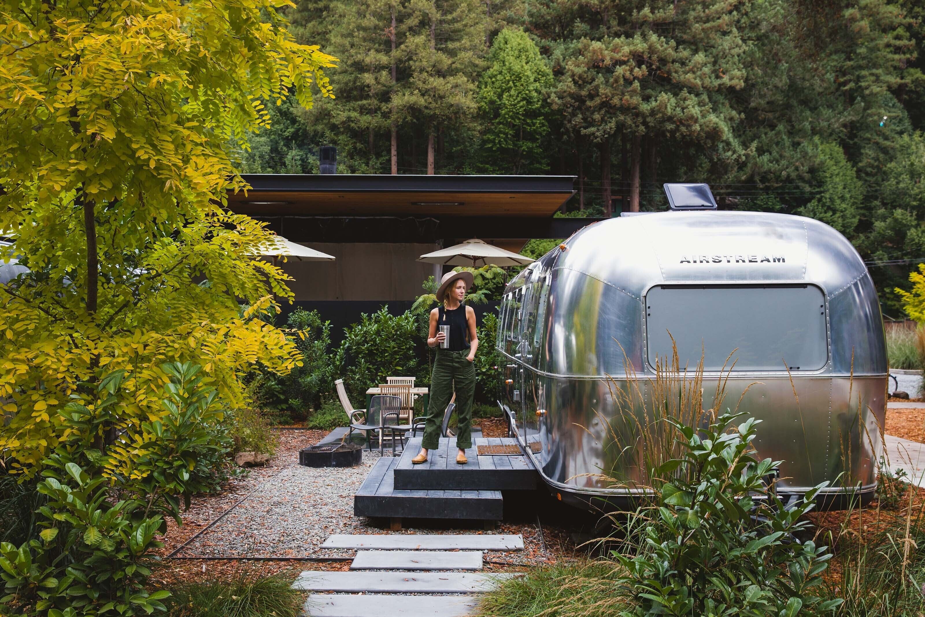 A person standing on a set of steps leading to a silver trailer labeled "AIRSTREAM" in a garden with trees and a modern building in the background.