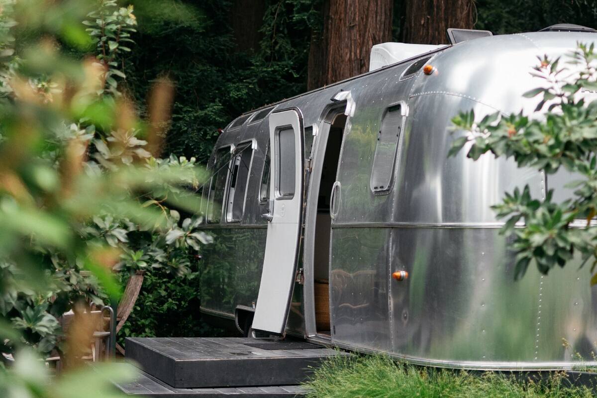A silver trailer with a ramp and a door, surrounded by greenery.
