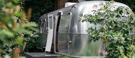 A silver trailer with a ramp and a door, surrounded by greenery.