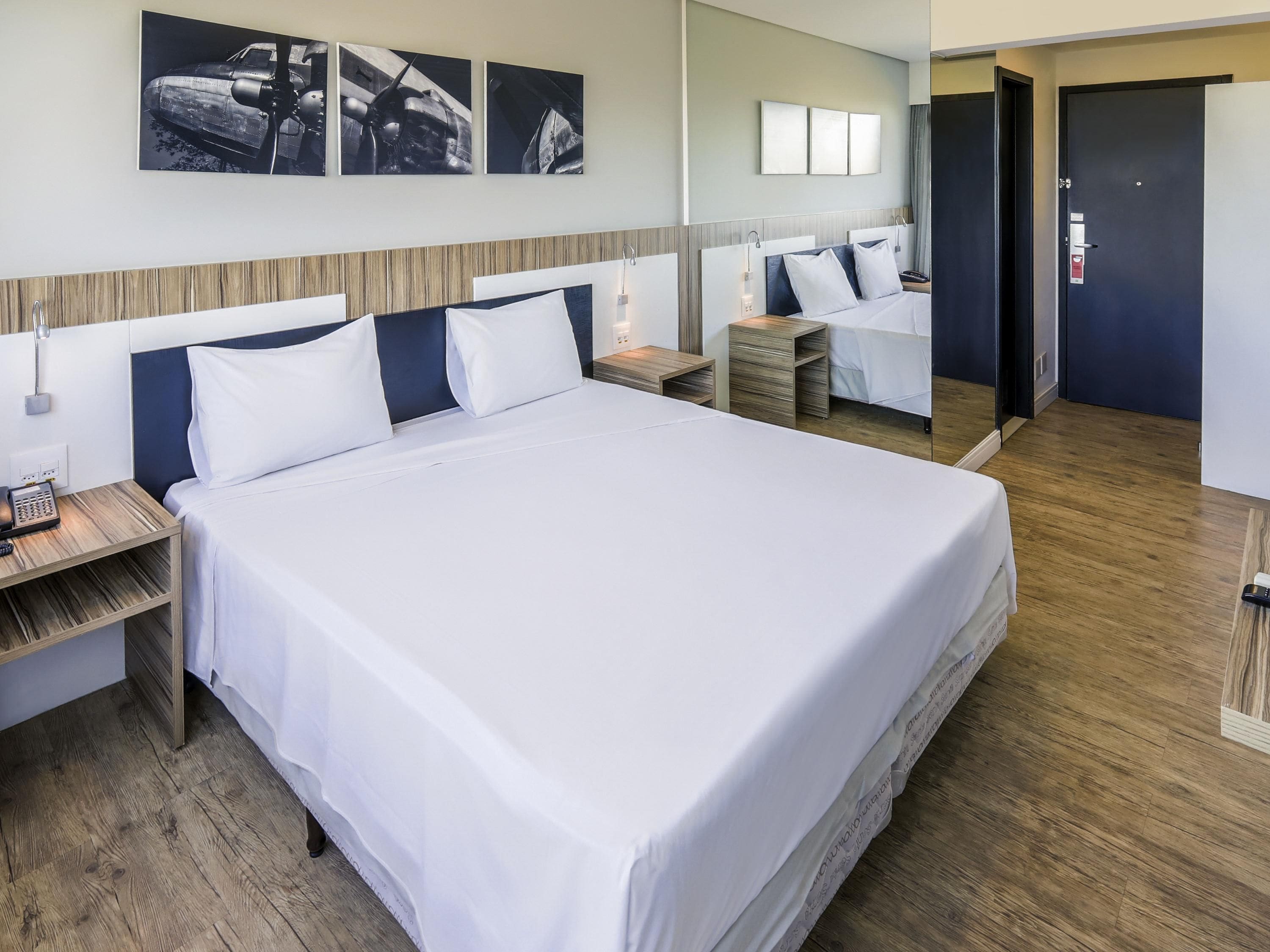 A hotel room with a large bed, bedside tables, a nightstand, a mirror, and artwork on the wall.