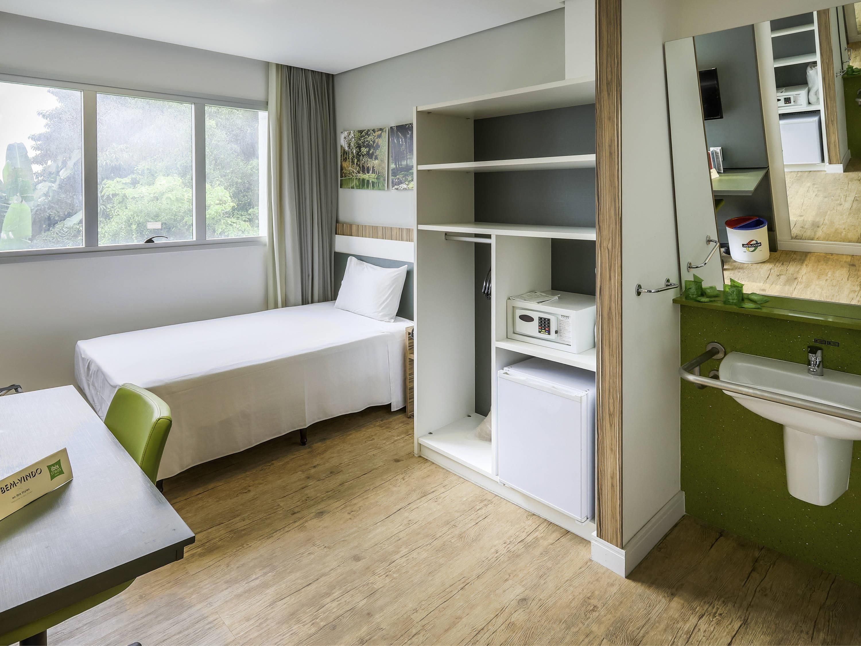 Quarto de hotel com uma cama, uma escrivaninha, uma estante com micro-ondas, pia e janela com cortinas.