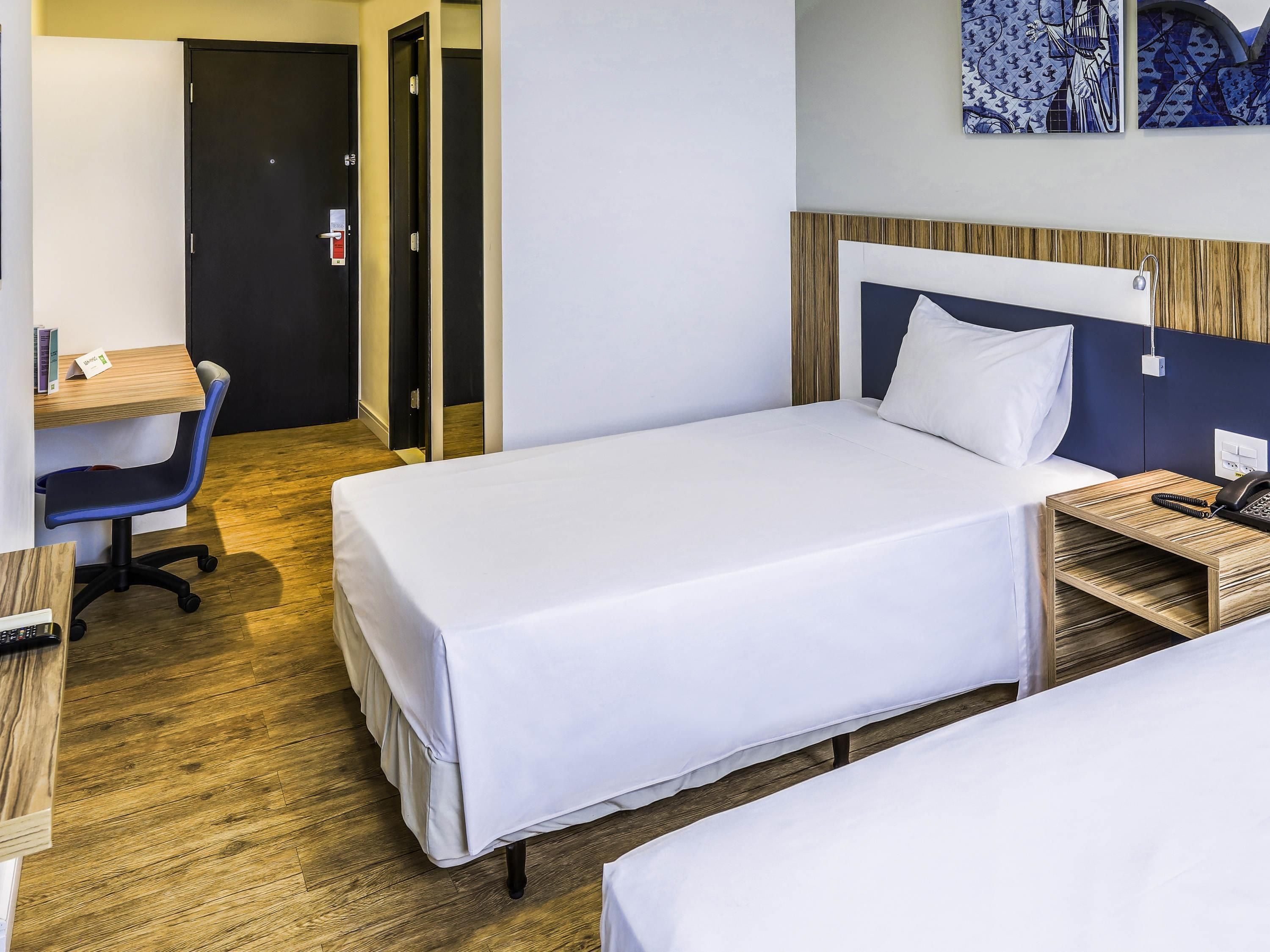 Quarto de hotel com duas camas de solteiro, uma escrivaninha com cadeira, telefone e obras de arte na parede.