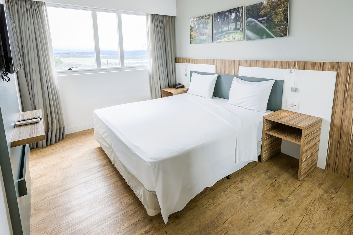 Habitación de hotel con una cama grande, mesitas de noche de madera, un escritorio y vistas al exterior a través de una ventana.