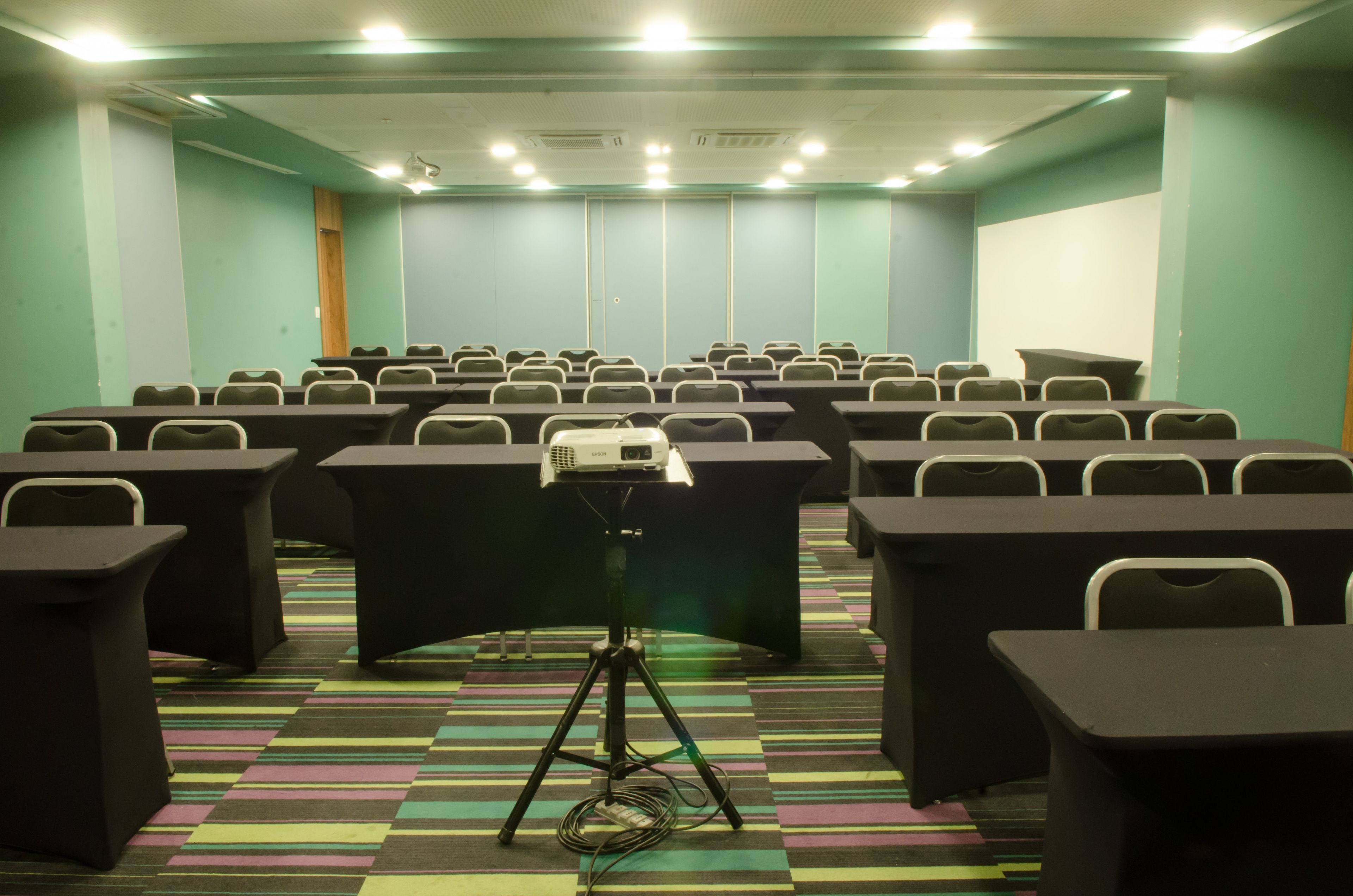 Une salle de conférence avec des rangées de tables et de chaises, un projecteur posé sur un trépied et un sol recouvert de moquette.
