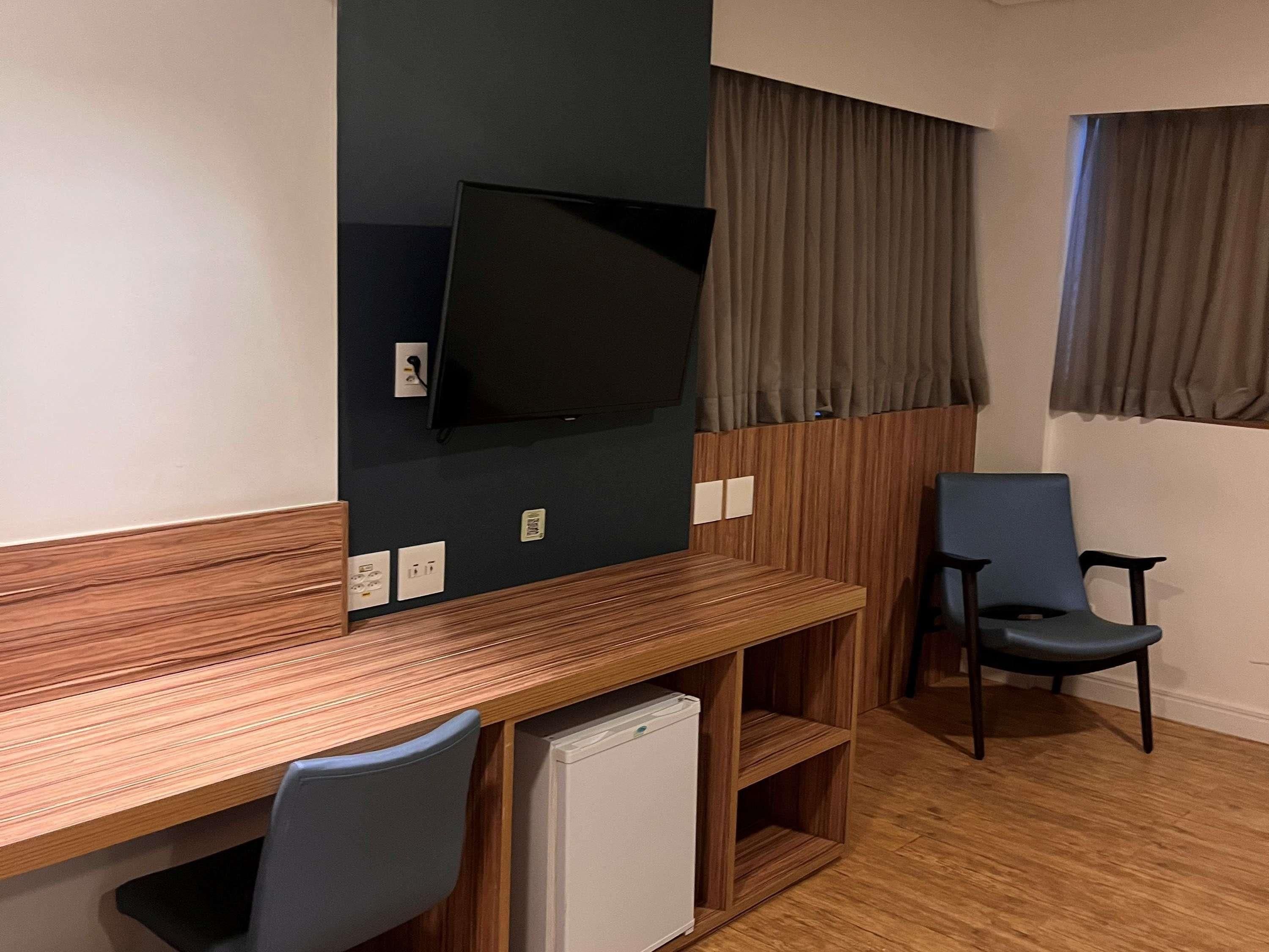 Una habitación con un escritorio de madera, un televisor montado en la pared, un refrigerador pequeño, una silla y una ventana con cortinas.
