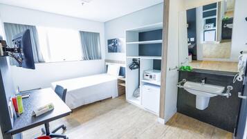 Quarto de hotel com cama, escrivaninha com cadeira, pequeno espaço de cozinha e banheiro com pia e espelho.