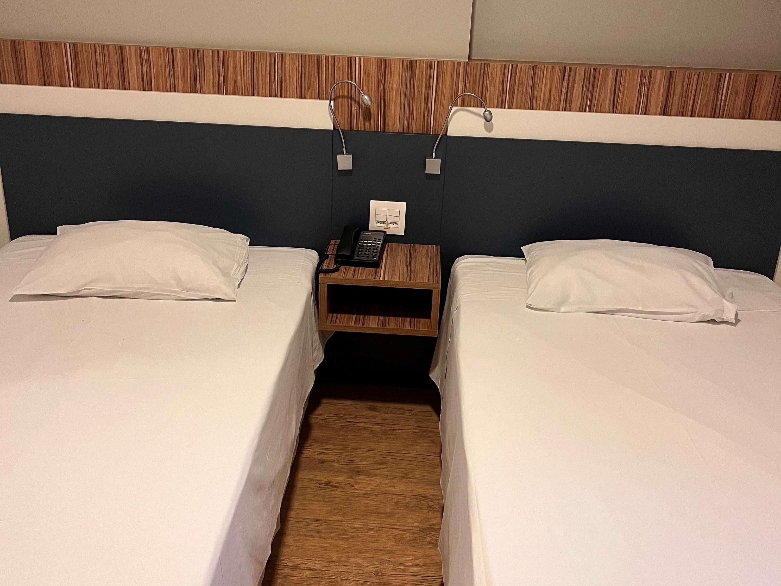 A hotel room with two single beds, a wooden nightstand with a phone, and a wall-mounted light fixture.