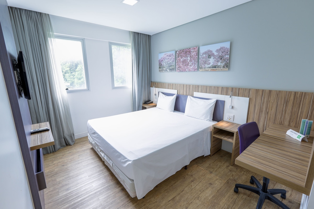 Una habitación de hotel moderna con una cama grande, un escritorio y una silla. Hay dos ventanas con cortinas, un televisor de pantalla plana montado en la pared y un estante pequeño con un control remoto.