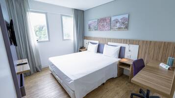 A modern hotel room with a large bed, a desk, and a chair. There are two windows with curtains, a flat-screen TV mounted on the wall, and a small shelf with a remote control.