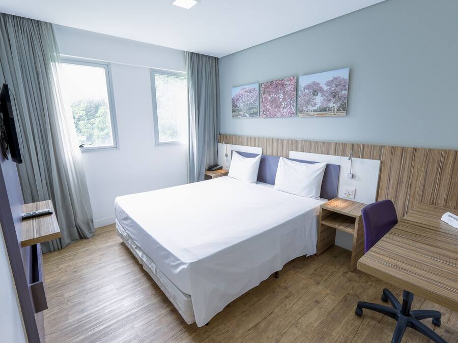 A modern hotel room with a large bed, a desk, and a chair. There are two windows with curtains, a flat-screen TV mounted on the wall, and a small shelf with a remote control.