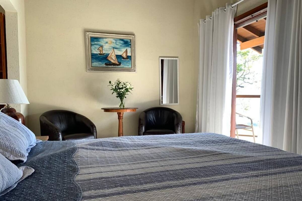 A bedroom with a large bed, two chairs, a small table with a plant, a painting, and a window with curtains.