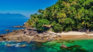 A tropical beach with clear turquoise waters, a rocky shoreline, and dense palm trees. - Pousada Irmaos do Mar (Ilhabela)