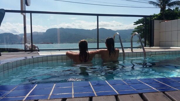 A swimming pool with two people in the water, surrounded by a wooden deck and a glass railing. - Pousada Irmaos do Mar (Ilhabela)