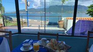 A table set with breakfast, overlooking a coastal view with a beach and mountains. - Pousada Irmaos do Mar (Ilhabela)