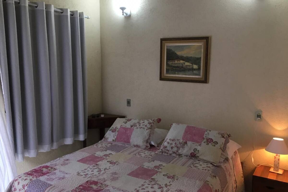 A bedroom with a bed, a nightstand, a lamp, a painting on the wall, and a window with curtains.