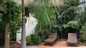 Un patio con banca de madera, abundante vegetación y una estructura de techo de paja.