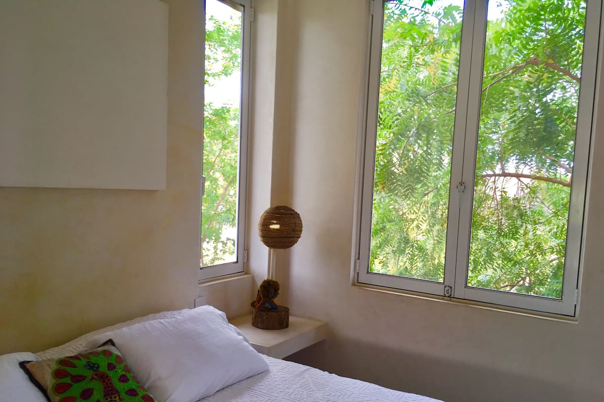 A bedroom with a bed, a window with a view of trees, and a small table with a lamp.