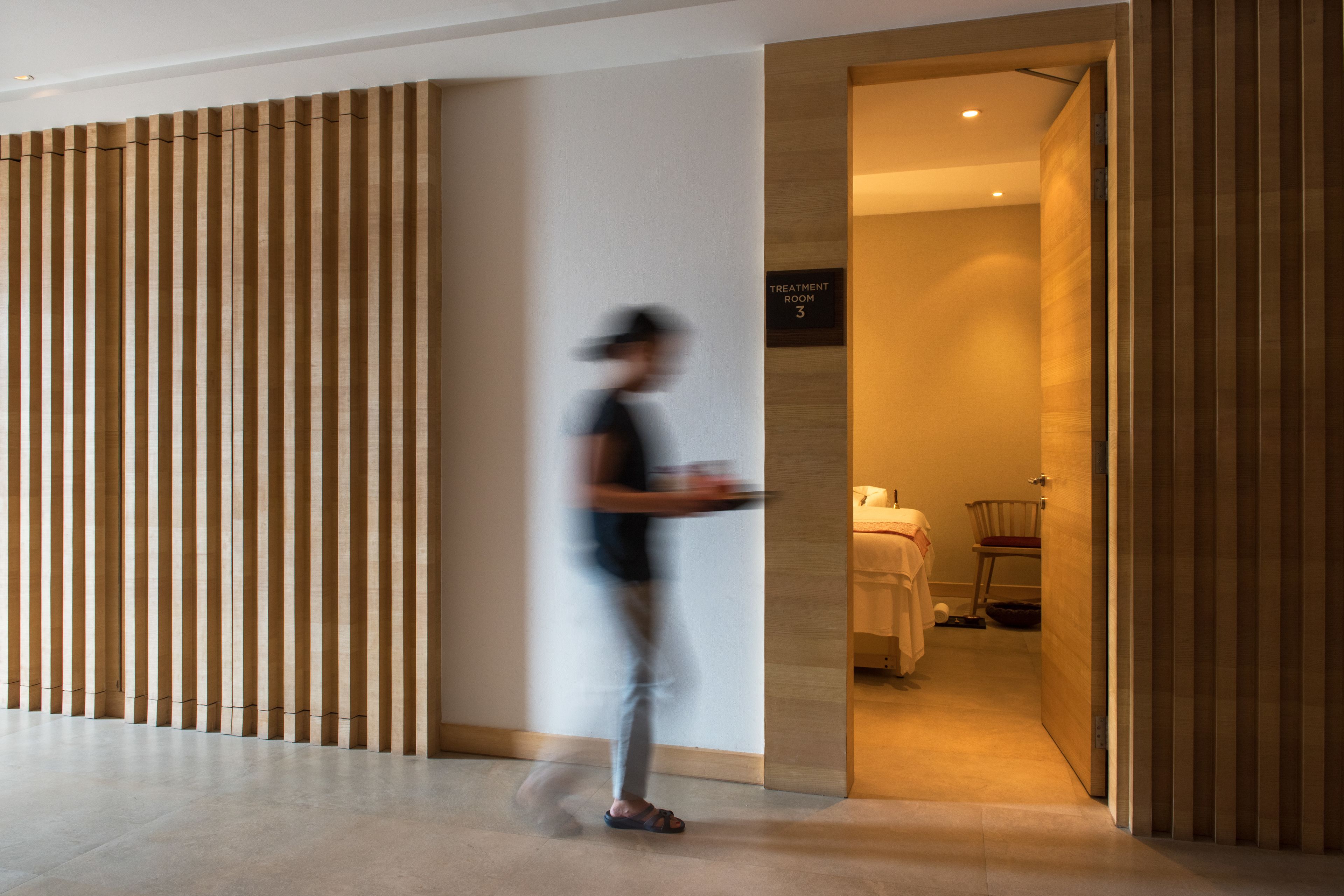 A modern hotel lobby with a wooden partition, a sign indicating 'Treatment Room 3', and a glimpse of a bed and chair in the background.