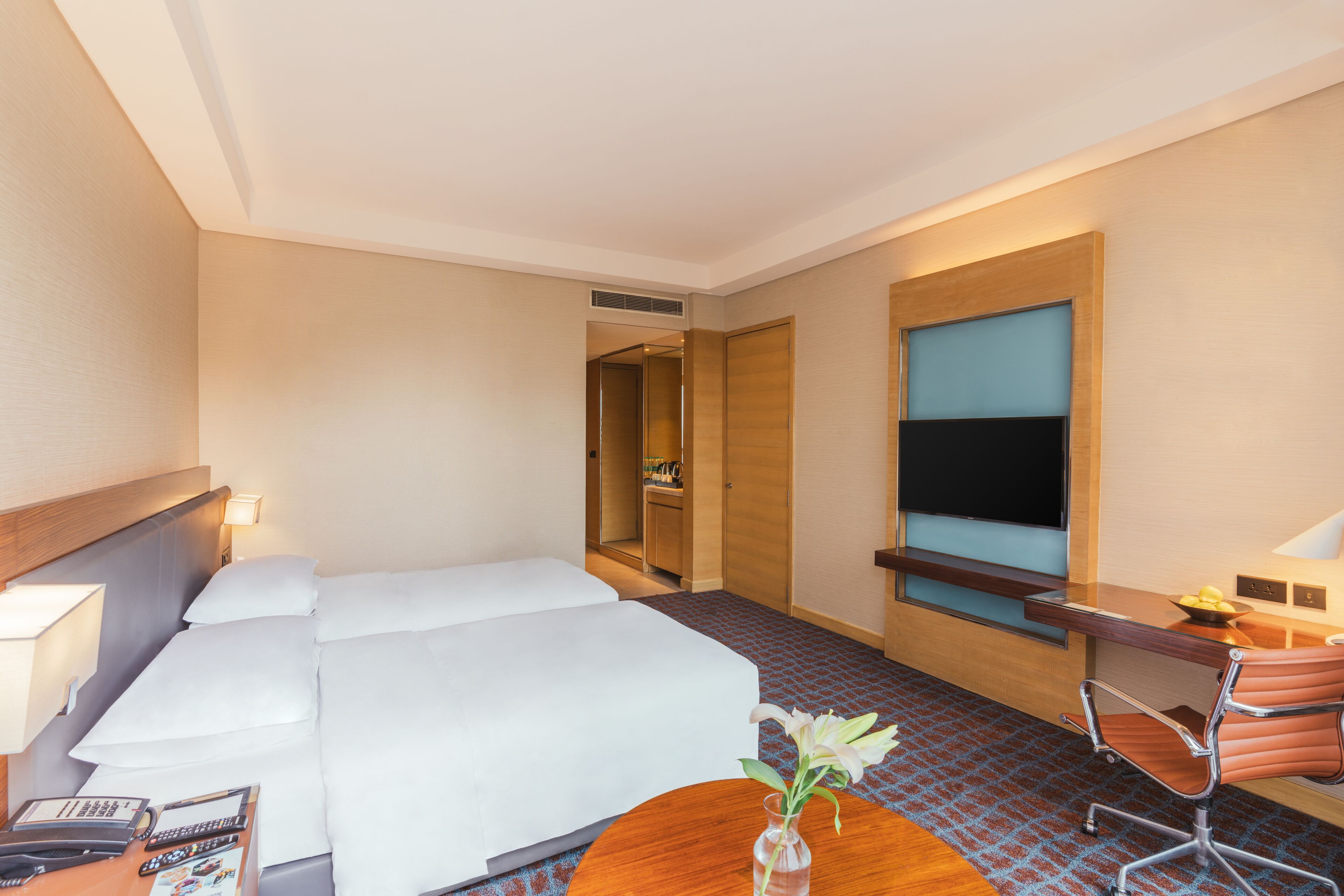 A hotel room with a large bed, a desk with a chair, a TV, and a small table with a vase of flowers.