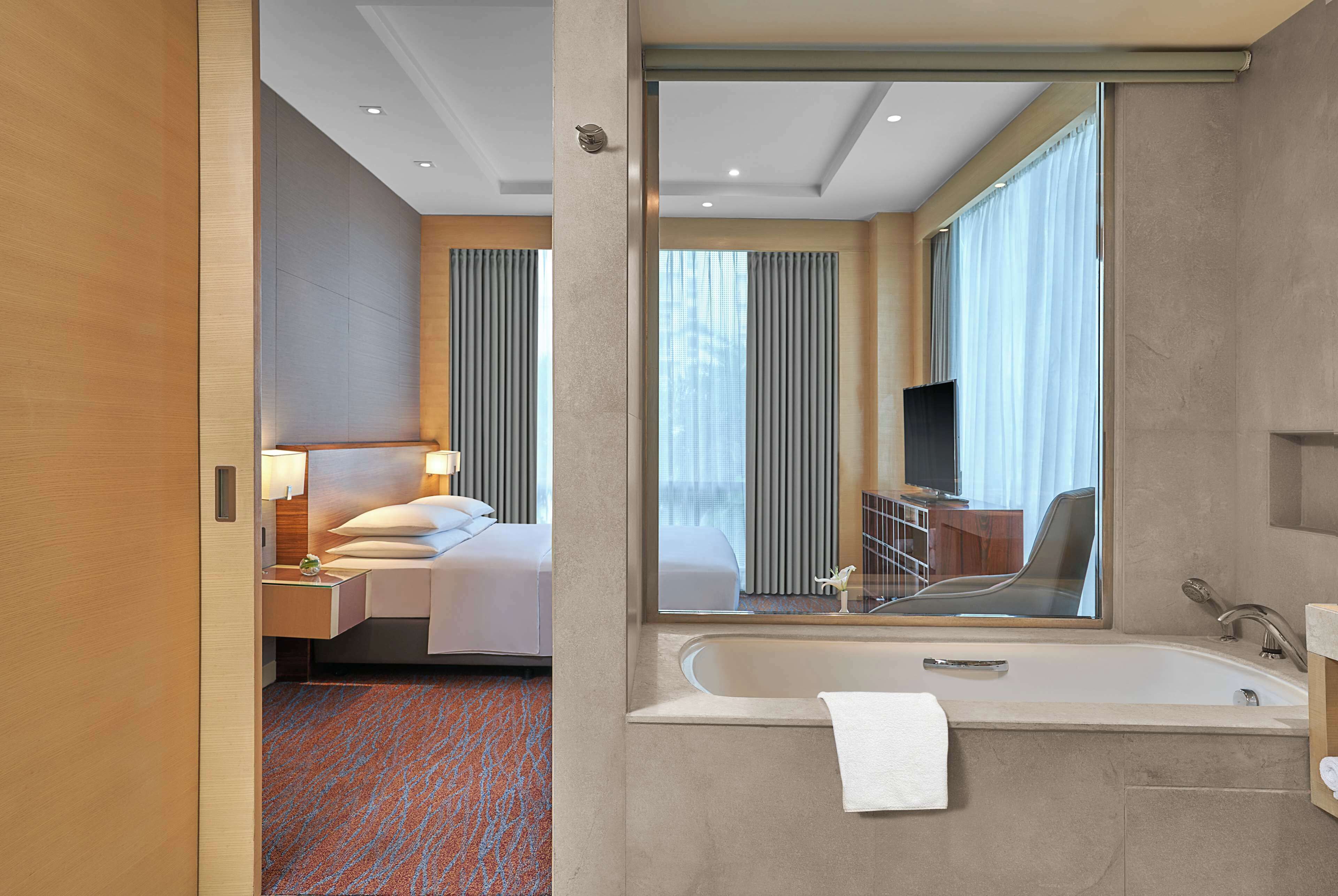 A modern hotel room with a large bed, a sitting area with a chair and table, a large mirror, a bathtub, and a television.